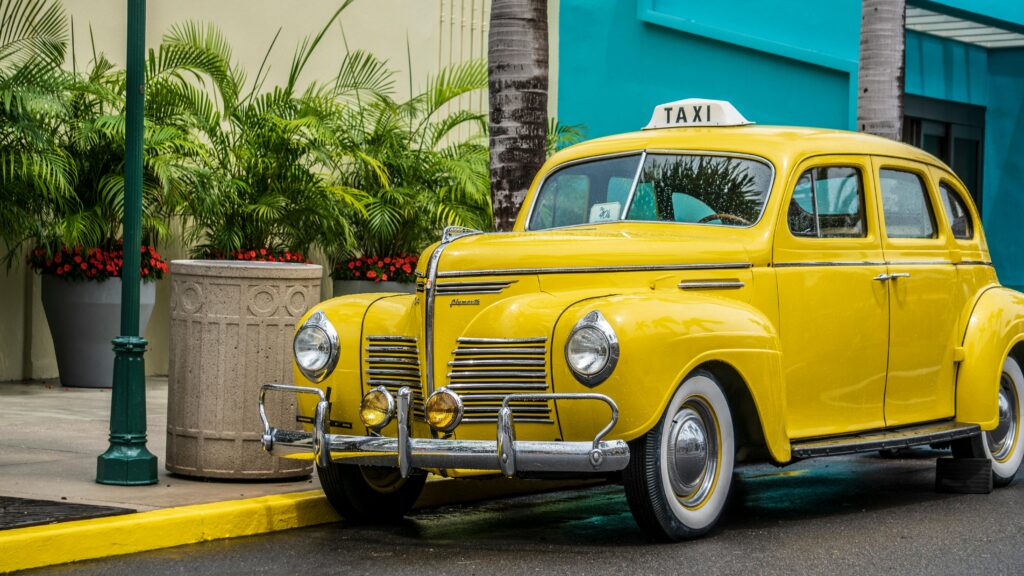 Vintage yellow taxi car parked on an urban street with lush green plants and palm trees.